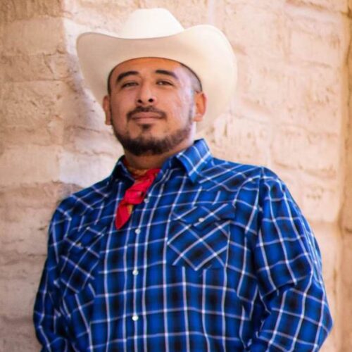 A picture of Joel Saldana Perez in a white cowboy hat, blue button up plaid shirt and a red handkerchief